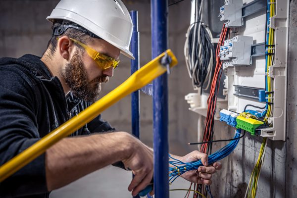 A male electrician works in a switchboard with an electrical connecting cable, connects the equipment with tools.